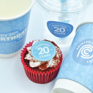 Two 12oz paper cups and a 12oz PET cup with a sticker celebrating the Printed Cup Company's 20th birthday. Both cups and the sticker are produced by the Printed Cup Company.